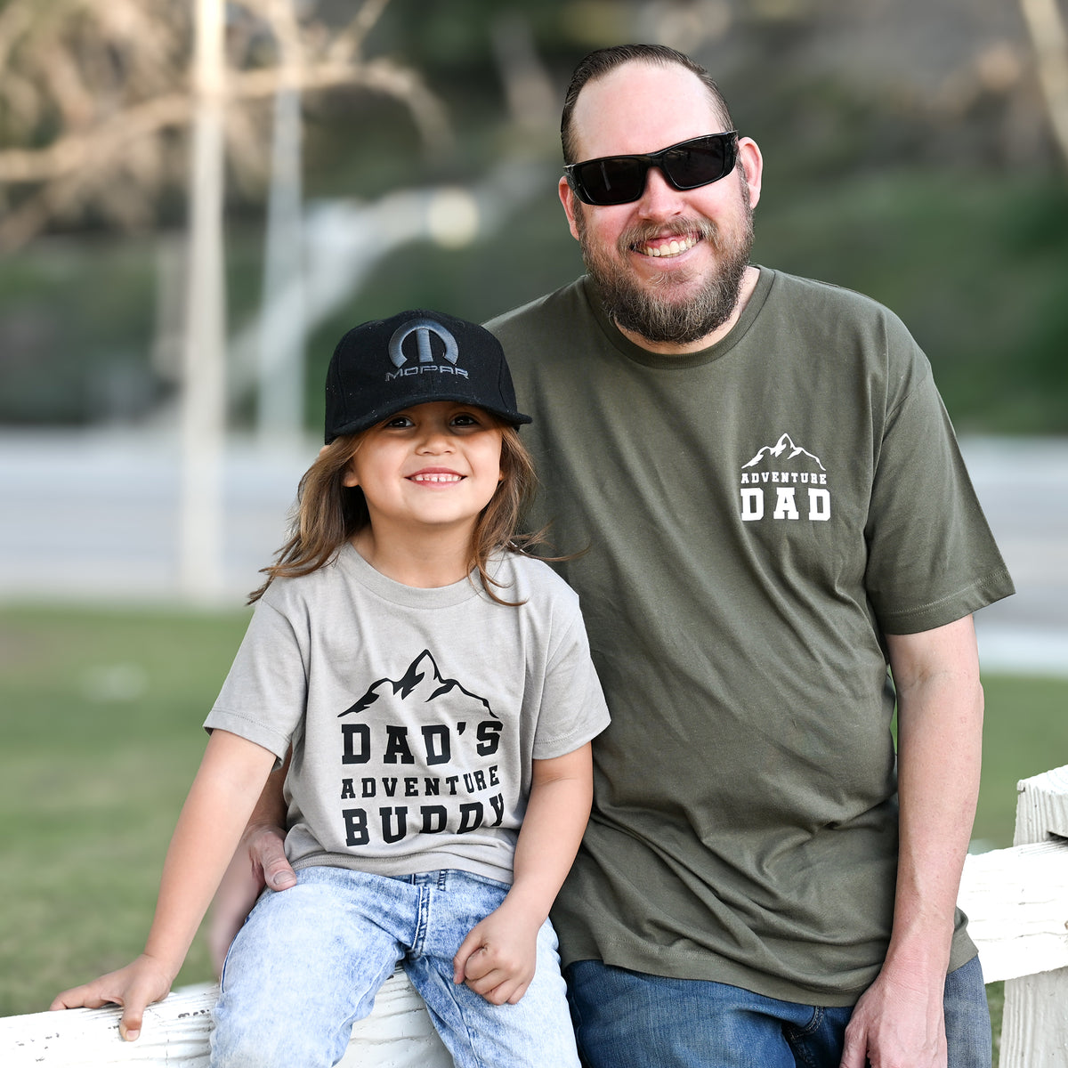 Man and child sitting together wearing matching t-shirts with 'DAD' and 'DAD'S ADVENTURE BUDDY' text.
