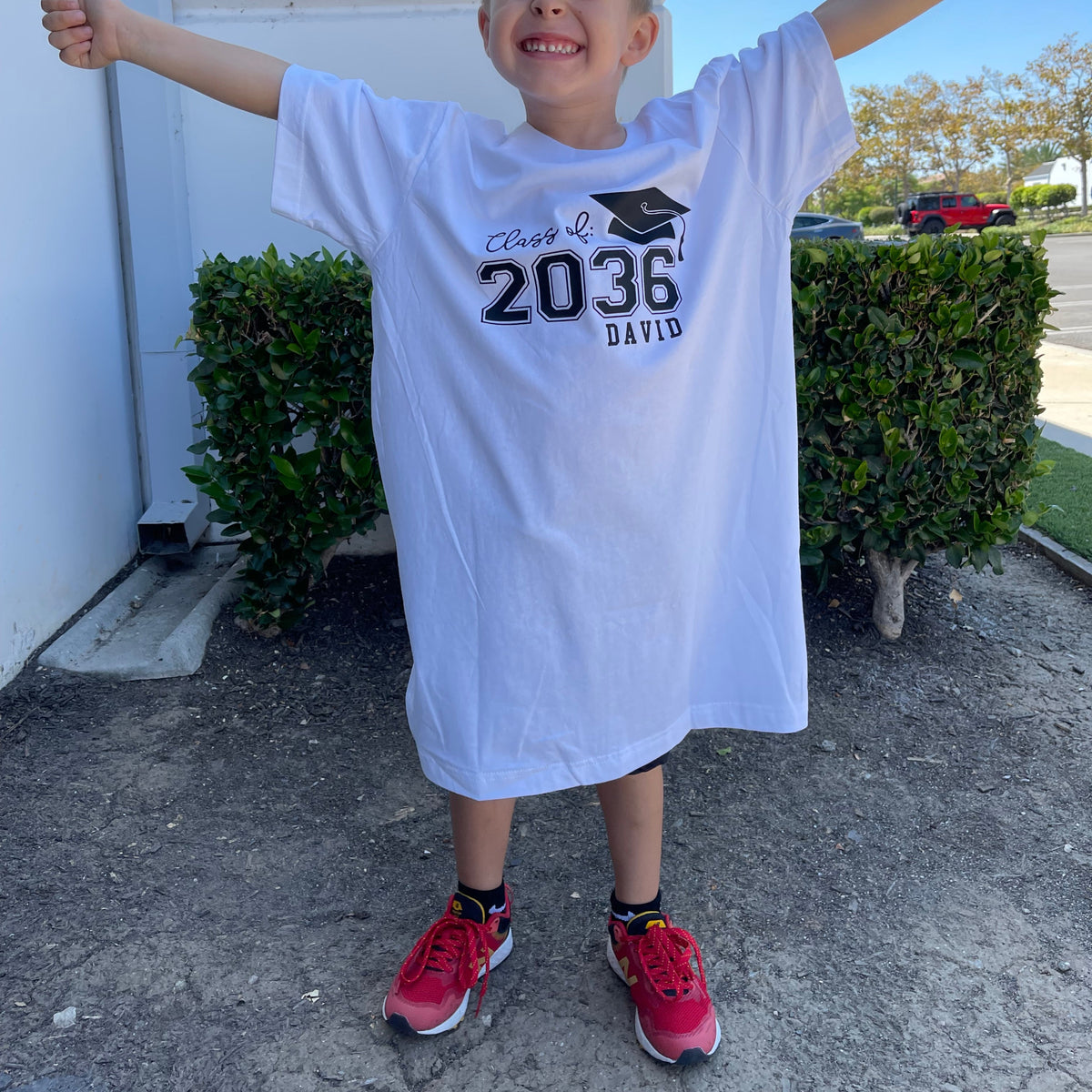 A child, dressed in a large white t-shirt featuring the school year and a custom name, stands outdoors.
