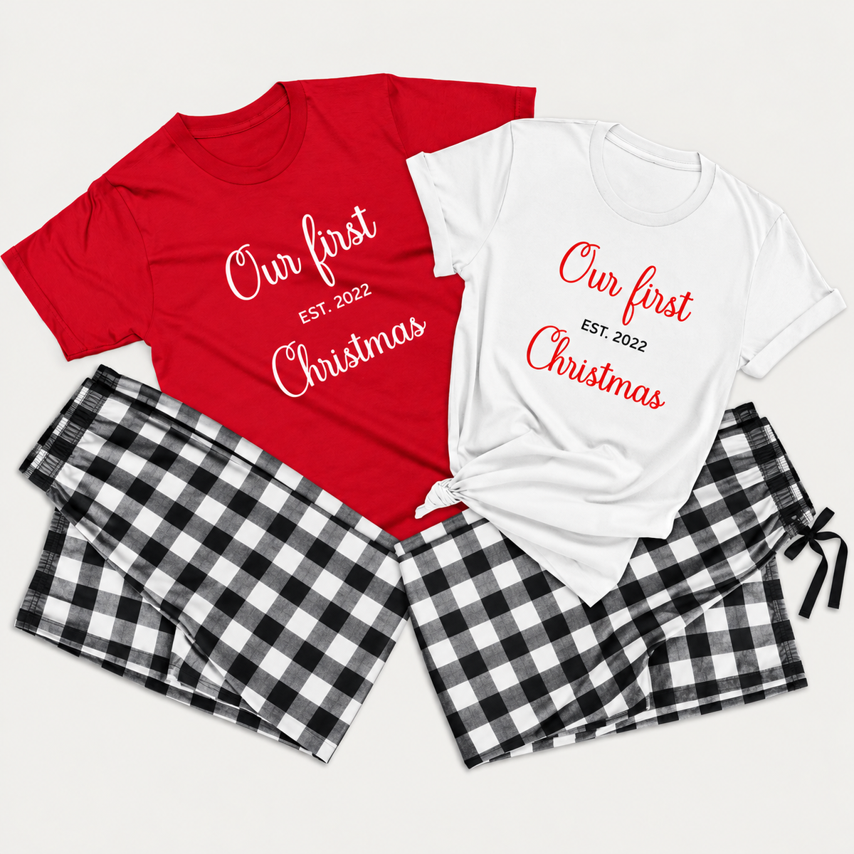 Matching Christmas pajamas featuring red and white T-shirts printed with “Our First Christmas” and “EST. 2022,” paired with black and white buffalo plaid flannel pajama pants in a clean studio flat lay.