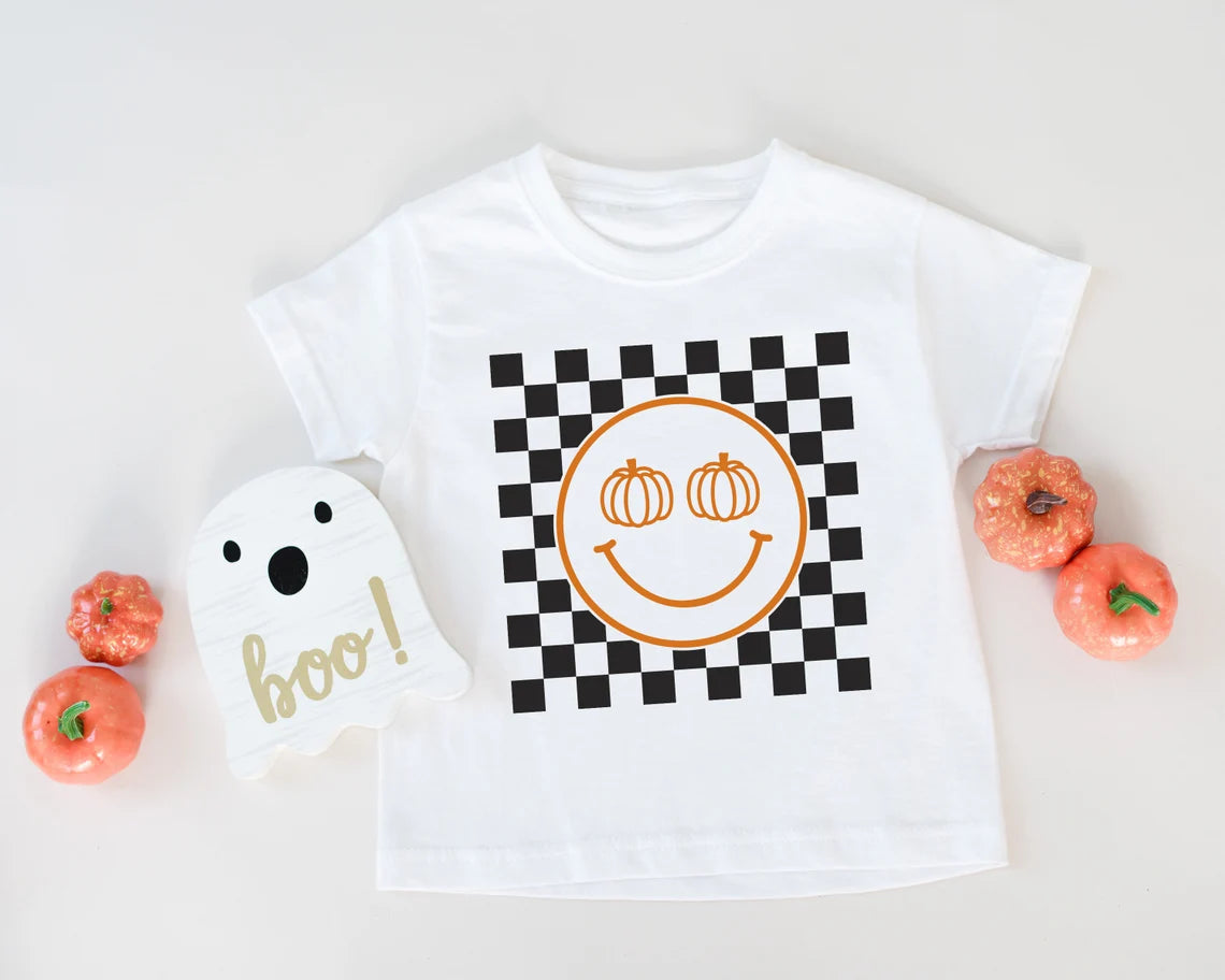 A white checkered toddler t-shirt with a happy smiley face and two pumpkin designs in orange, placed on a surface with checkered pattern, next to a 'BOO!' sign and some candy corn.