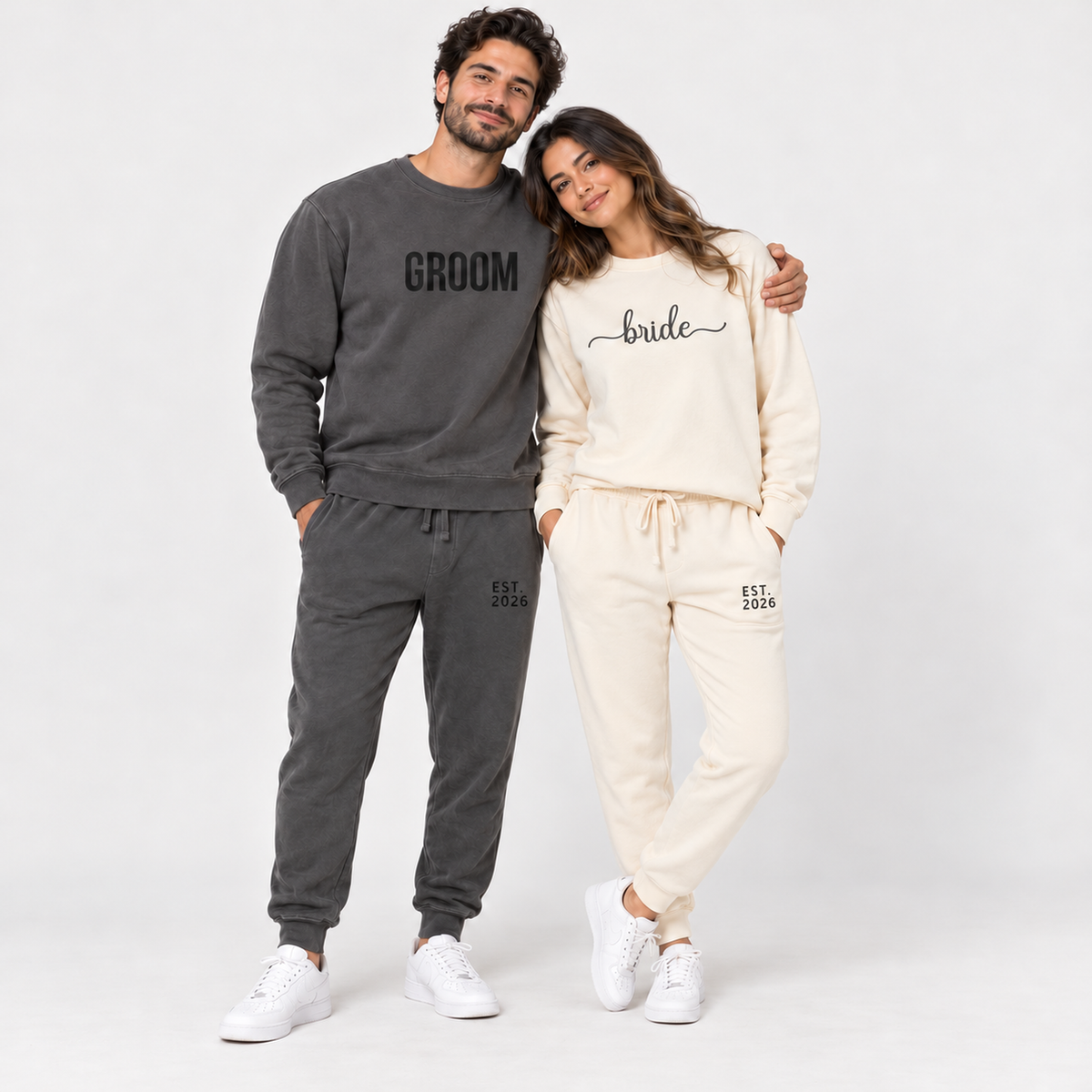 Young couple wearing matching bride and groom sweatshirts and joggers with EST. 2026 wedding date prints, studio portrait