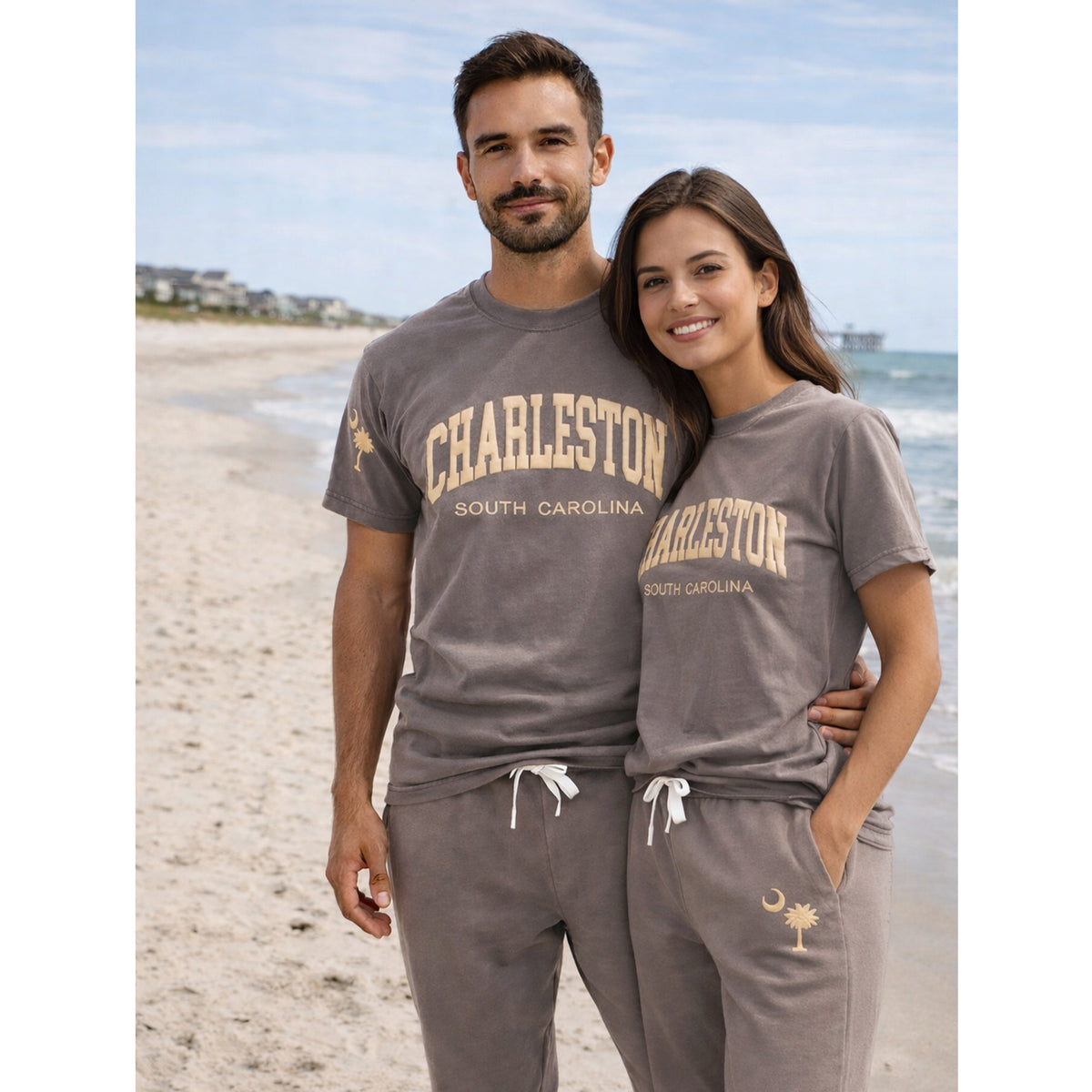 Couple wearing matching Charleston South Carolina Comfort Colors t-shirts and sweatpants with puff print and palm tree crescent moon logo on beach, unisex lounge set.