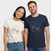 Smiling couple wearing white and navy matching T-shirts with pinky-promise hand illustrations and “Since 2018” text.
