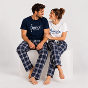 Engaged couple sitting together on a white studio cube, smiling at each other while wearing matching fiancé and fiancée T-shirts with coordinating navy and silver plaid flannel pajama pants.
