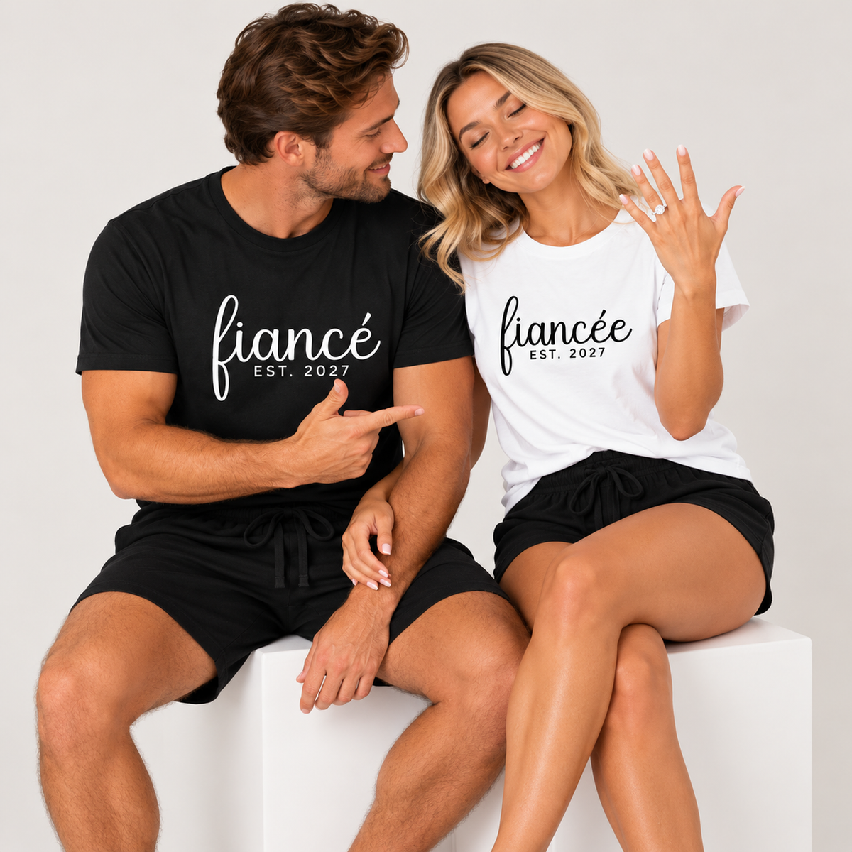 Engaged couple wearing personalized fiancé and fiancée matching pajama T-shirts and shorts featuring EST. 2027, sitting on a white studio cube as the man points to her engagement ring.
