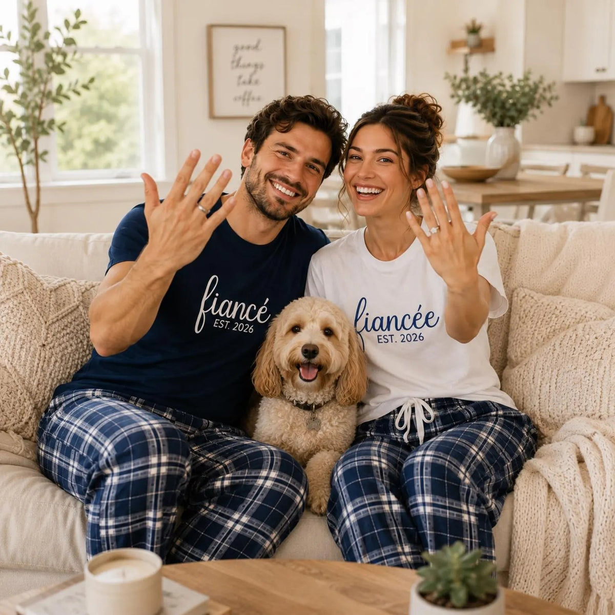 Engaged couple wearing matching fiancé and fiancée T-shirts with coordinating flannel pajama pants, showing their engagement rings while sitting on a sofa with their dog.