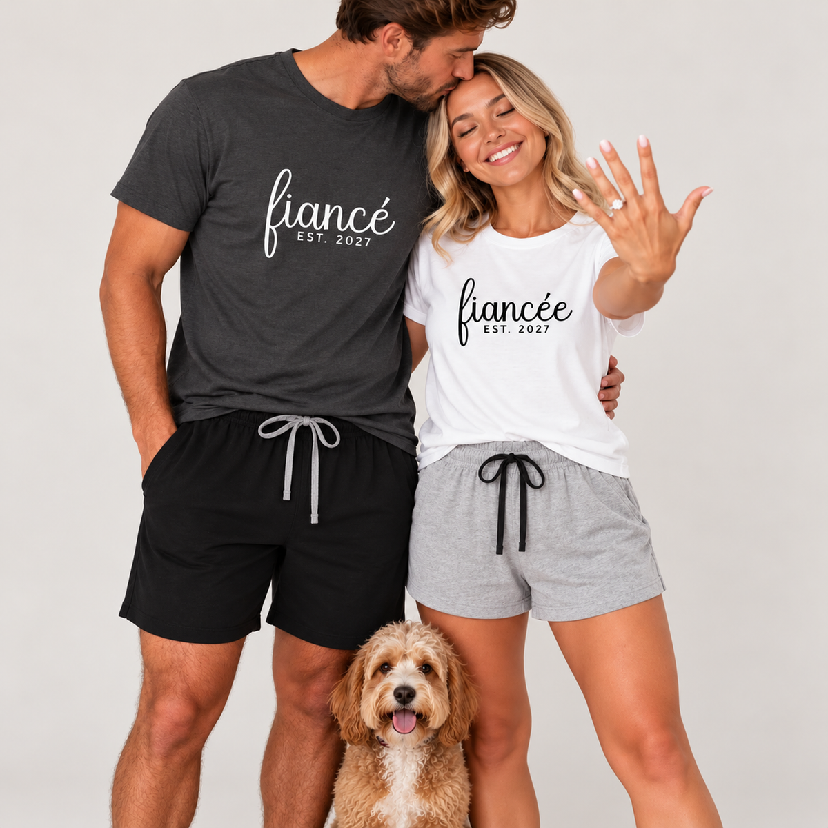 Engaged couple wearing personalized fiancé and fiancée matching pajama T-shirts and shorts featuring EST. 2027, with the man in an asphalt T-shirt and black shorts and the woman in a white T-shirt and athletic heather shorts, posing with their dog in a studio.