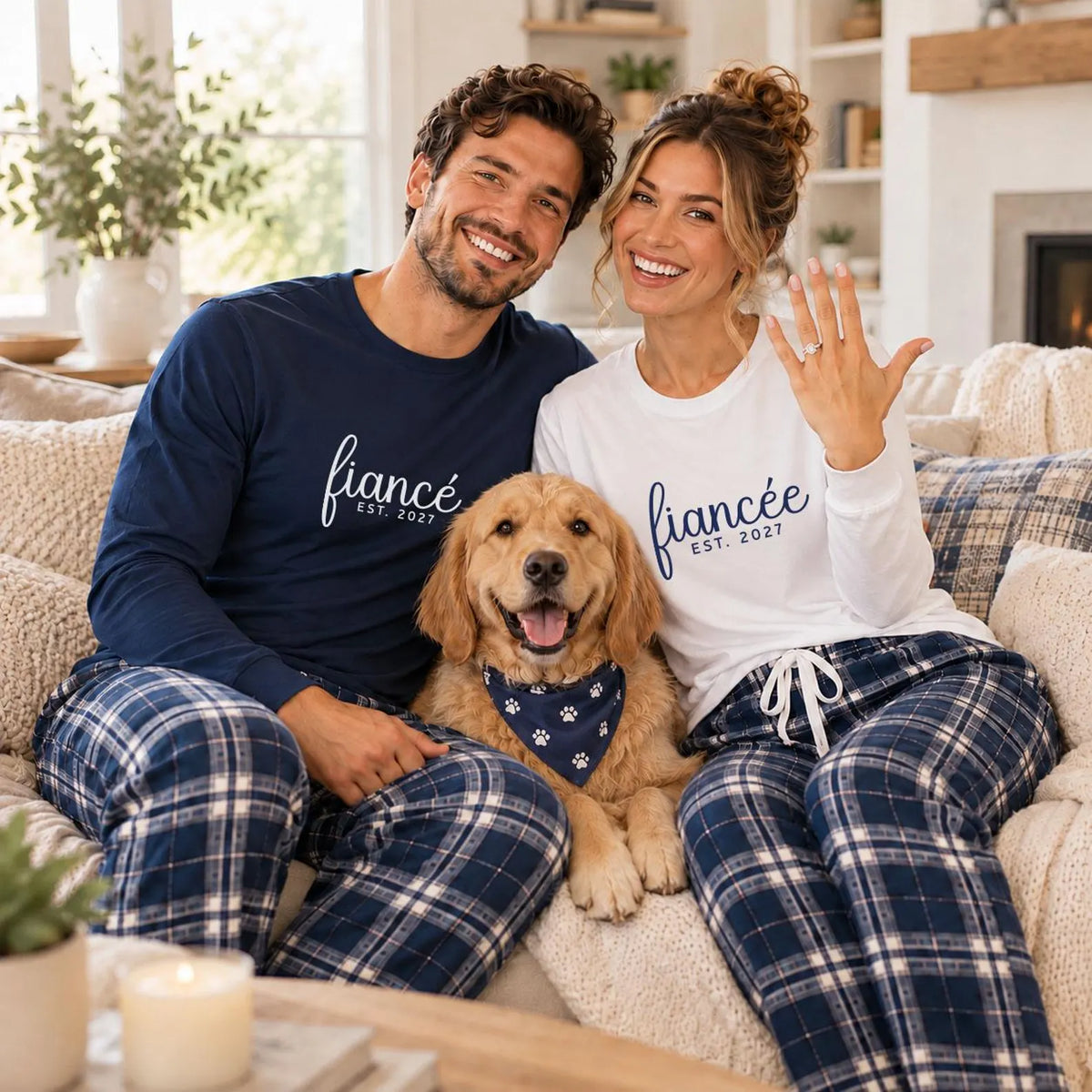 Engaged couple wearing matching fiancé and fiancée long sleeve tees and plaid flannel pajama pants, sitting on a sofa with their dog.