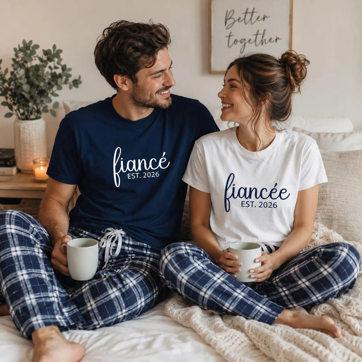 Engaged couple wearing matching fiancé and fiancée unisex T-shirts with coordinating navy and silver plaid flannel pajama pants, enjoying coffee together while sitting on a bed.