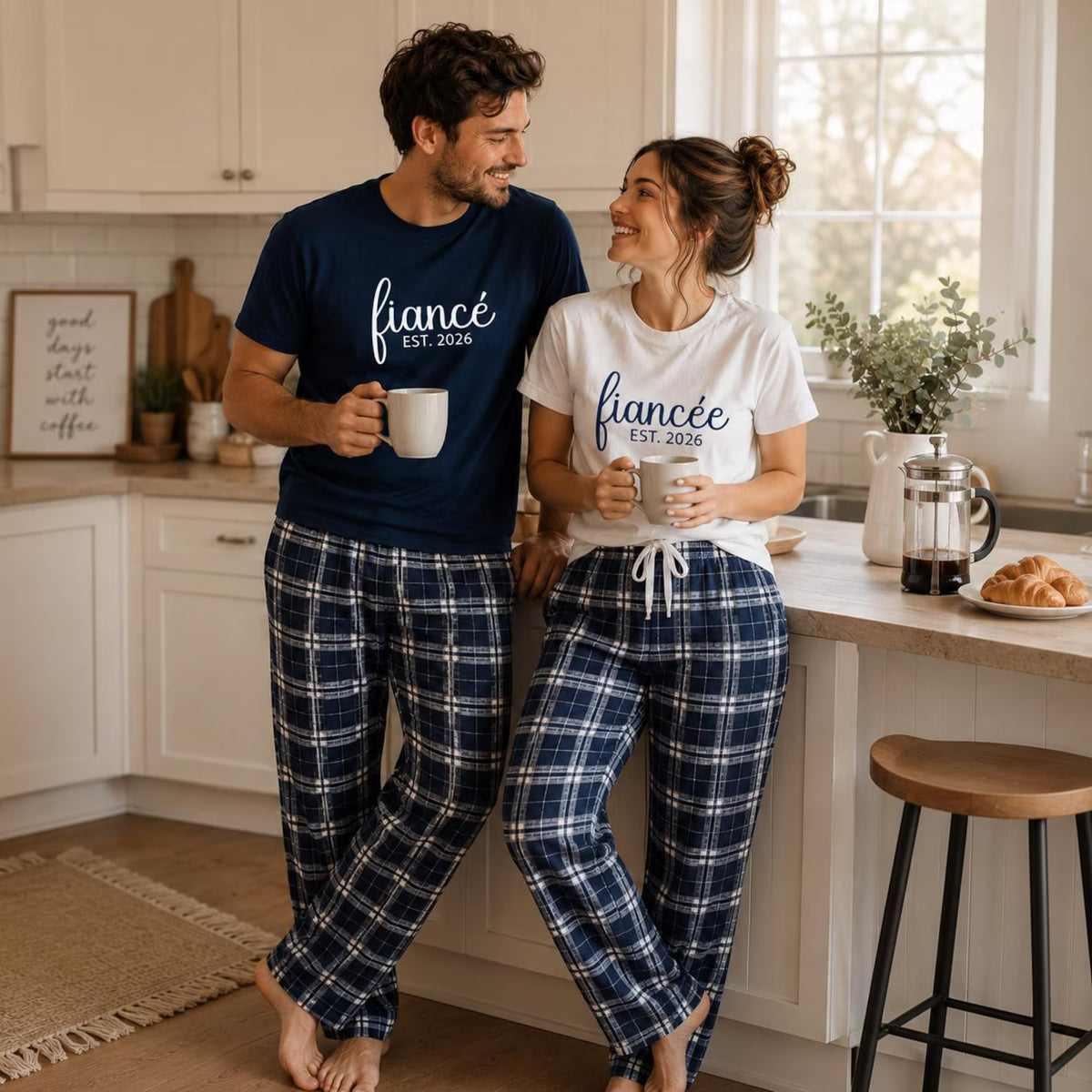 Engaged couple wearing matching fiancé and fiancée T-shirts in unisex styles with navy and silver plaid flannel pajama pants, enjoying coffee together in the kitchen.
