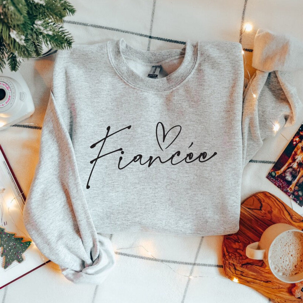 Athletic Heather Fiancée sweatshirt featuring elegant script lettering and a heart design, styled in a festive Christmas flat lay with holiday decorations, a book, and a cup of coffee.