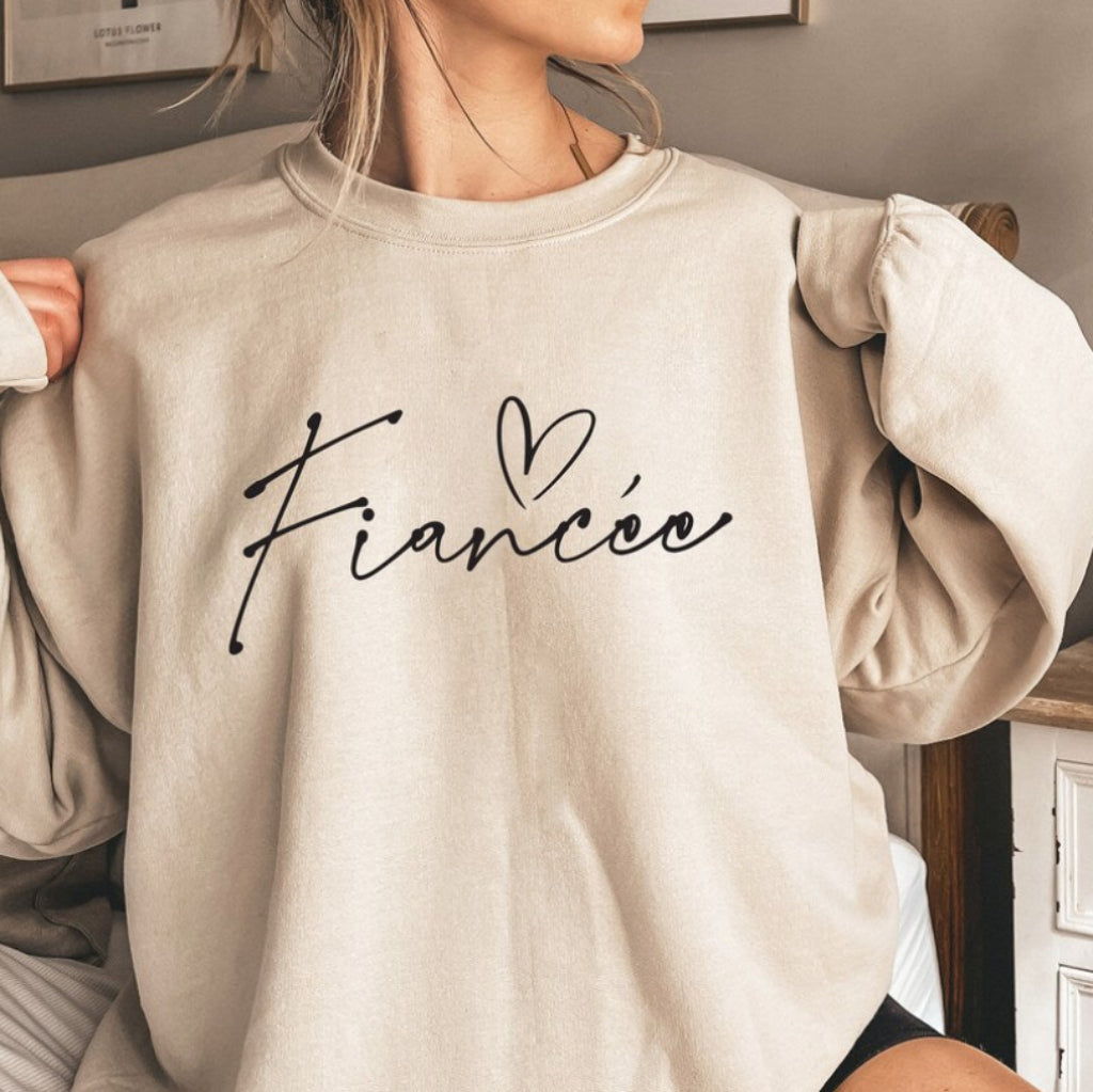 Woman wearing a Sandstone Fiancée sweatshirt featuring elegant script lettering and a heart design on the front.