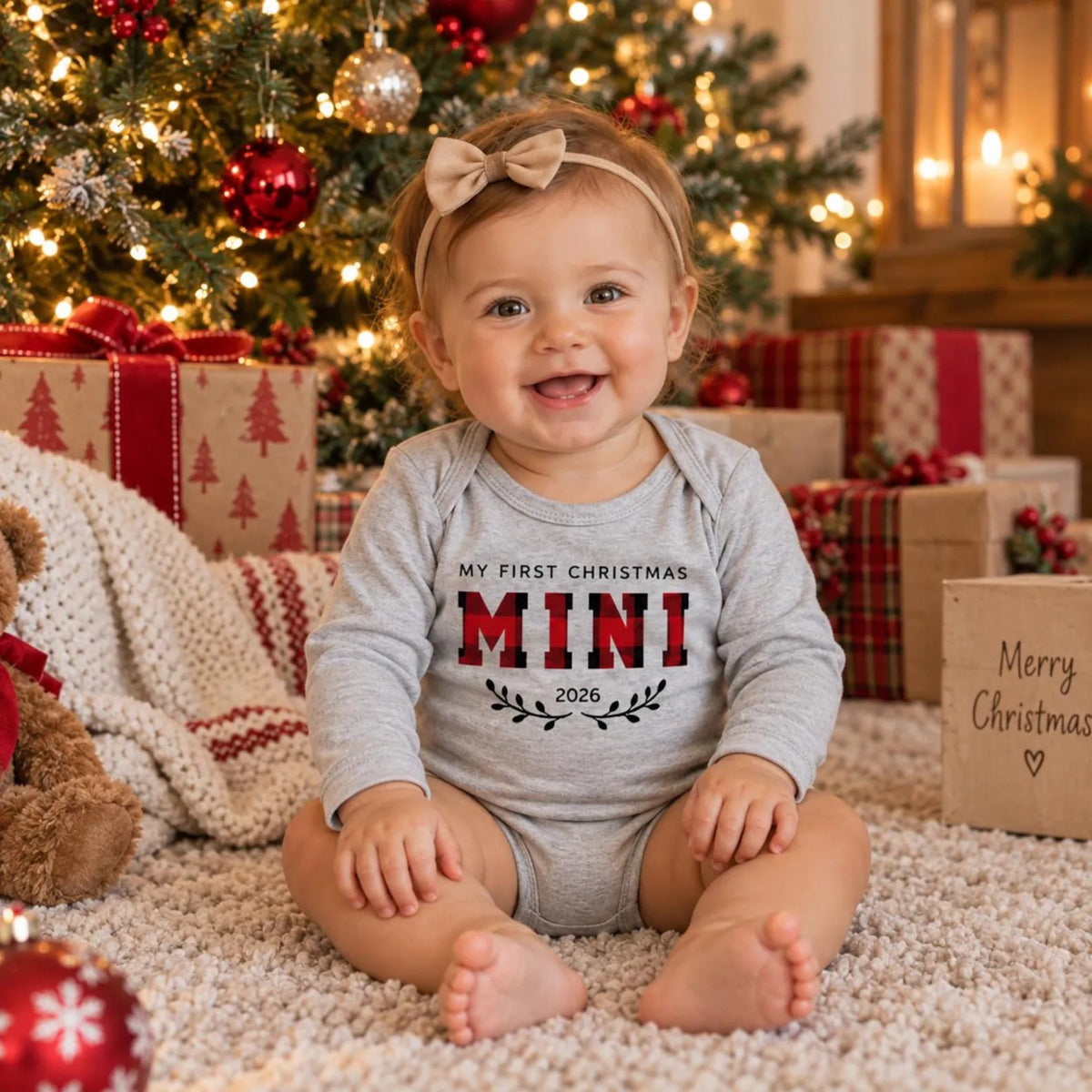 Smiling baby wearing a personalized First Christmas Mini 2026 gray long-sleeve bodysuit with a red and black buffalo plaid design, sitting on a cozy rug in a festive Christmas living room.