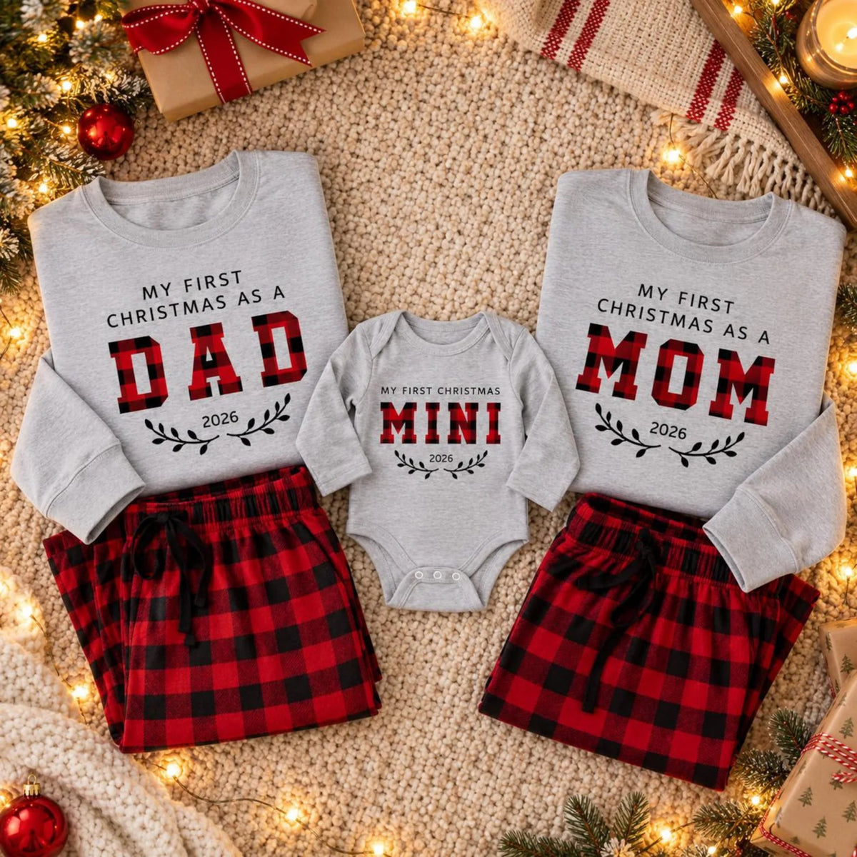Personalized matching First Christmas 2026 family outfits featuring Dad, Mom, and Mini designs on sweatshirts and a baby bodysuit, paired with red and black buffalo plaid pajama pants, styled on a cozy knitted blanket with festive Christmas decorations.