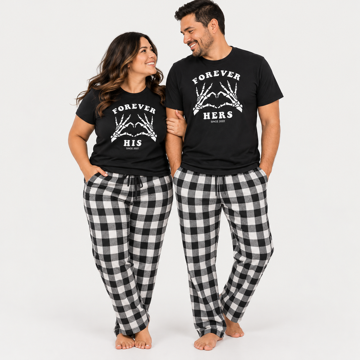 Couple wearing matching black Forever His and Forever Hers T-shirts with black and white flannel pajama pants in a studio setting