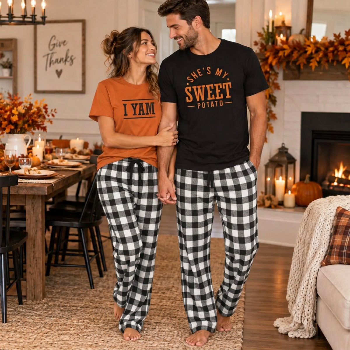 A couple wearing funny Thanksgiving matching T-shirts, with the man in a black She's My Sweet Potato T-shirt and the woman in an autumn She's My I Yam T-shirt, paired with white-and-black plaid flannel pajama pants.