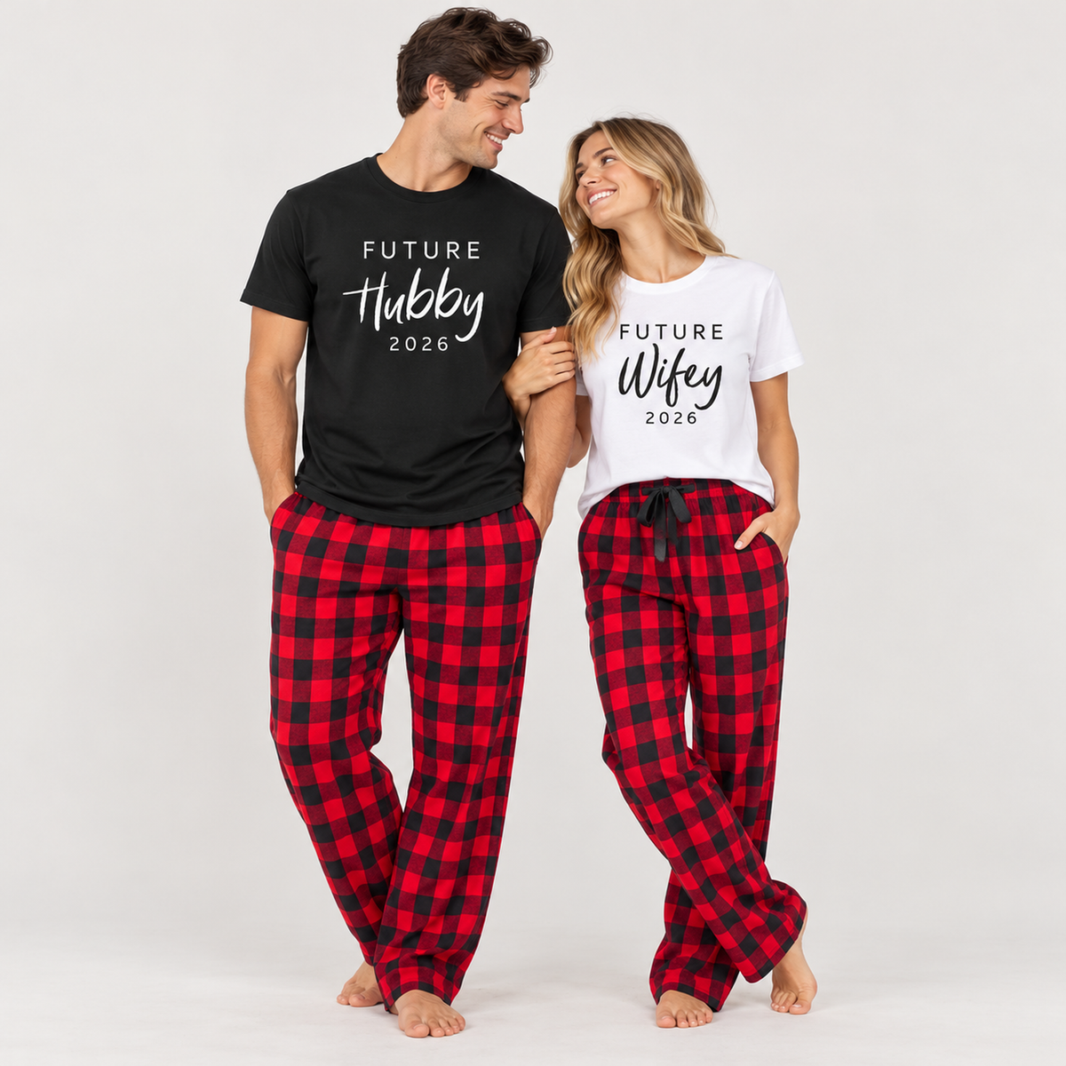 Young couple wearing matching Future Hubby and Future Wifey T-shirts with 2026 prints and red-and-black buffalo plaid flannel pajama pants in a clean studio setting.