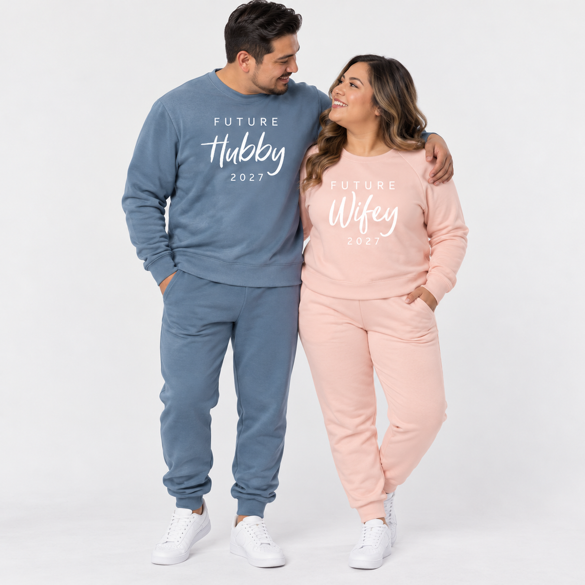 A middle-aged couple wearing matching Pigment Slate Blue and blush pink sweatshirts and joggers featuring the original Future Hubby 2027 and Future Wifey 2027 prints, posing together in a clean studio.