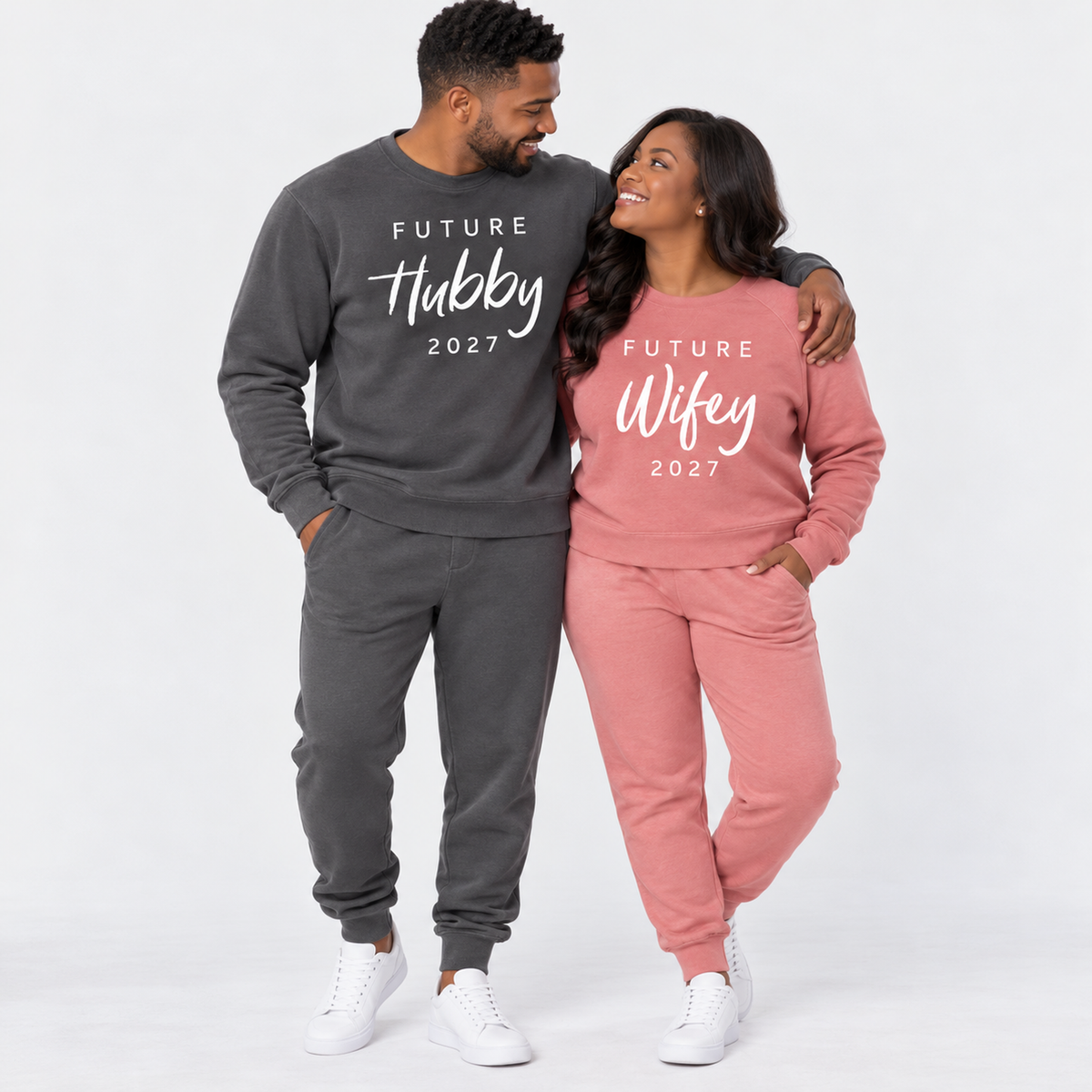 A couple wearing matching pigment black and dusty rose sweatshirts and joggers featuring Future Hubby 2027 and Future Wifey 2027 prints, posing together in a clean studio.