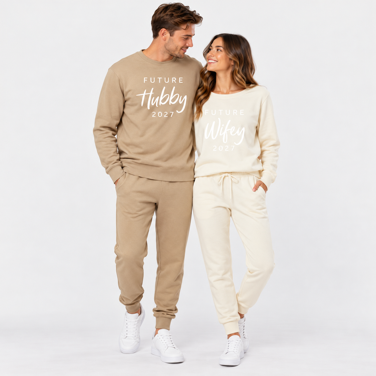 A couple wearing matching sandstone and bone sweatshirts and joggers printed with Future Hubby 2027 and Future Wifey 2027, posing together in a clean studio setting.