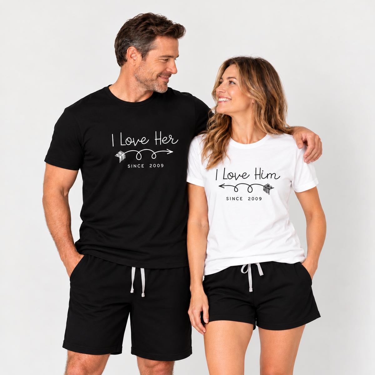Couple wearing matching I Love Her and I Love Him graphic T-shirts with black shorts in a clean studio setting.