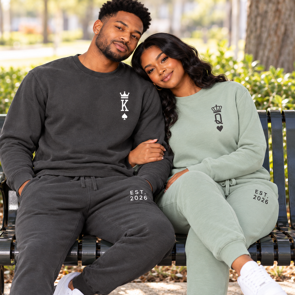 Couple wearing matching King and Queen sweatshirts and joggers with crown, playing card symbols, and EST. 2026 prints, seated together on a park bench