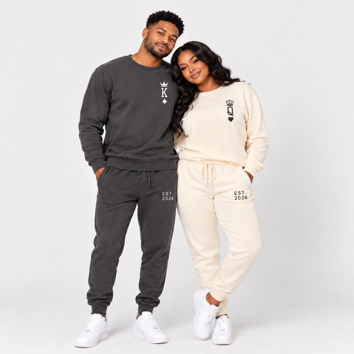 Couple wearing matching King and Queen sweatshirts and joggers featuring crown, playing card symbols, and EST. 2026 personalized prints in a studio portrait