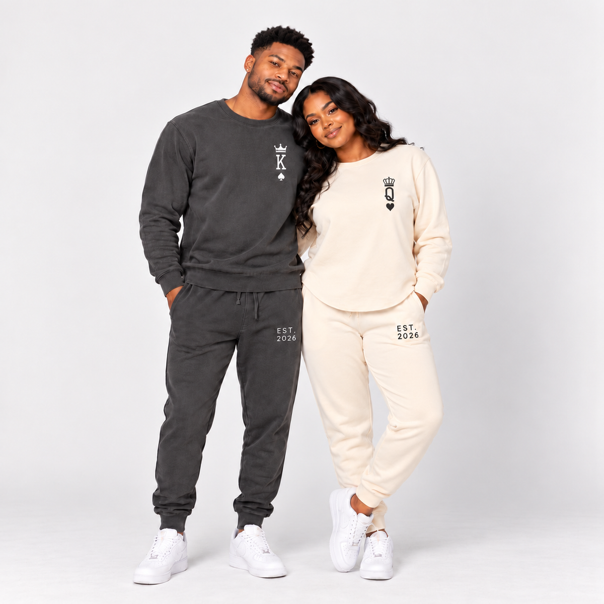 Couple wearing matching King and Queen sweatshirts and joggers with crown, playing-card symbols, and EST. 2026 personalization in a studio setting.