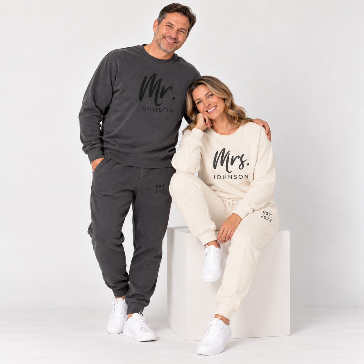 A couple wearing matching pigment black and bone sweatsuits personalized with Mr. Johnson and Mrs. Johnson lettering and EST. 2022 details, posing together in a clean studio.