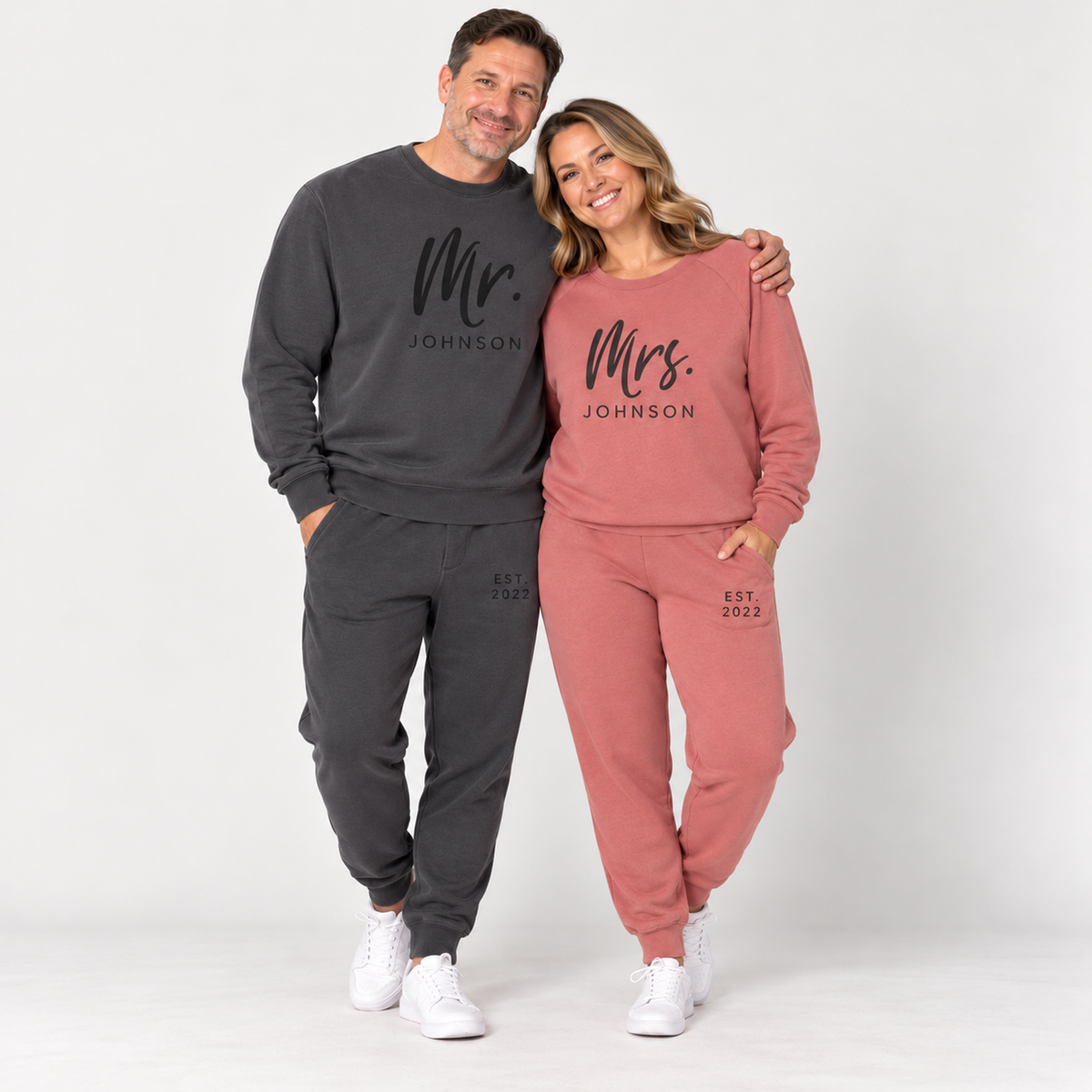A couple wearing matching pigment black and dusty rose sweatsuits personalized with Mr. Johnson and Mrs. Johnson lettering and EST. 2022 details, posing together in a clean studio.