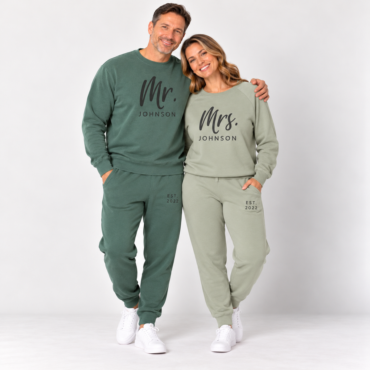 A couple standing together in matching pigment alpine green and sage sweatshirts and joggers personalized with Mr. Johnson and Mrs. Johnson lettering and EST. 2022 details, in a clean studio setting.