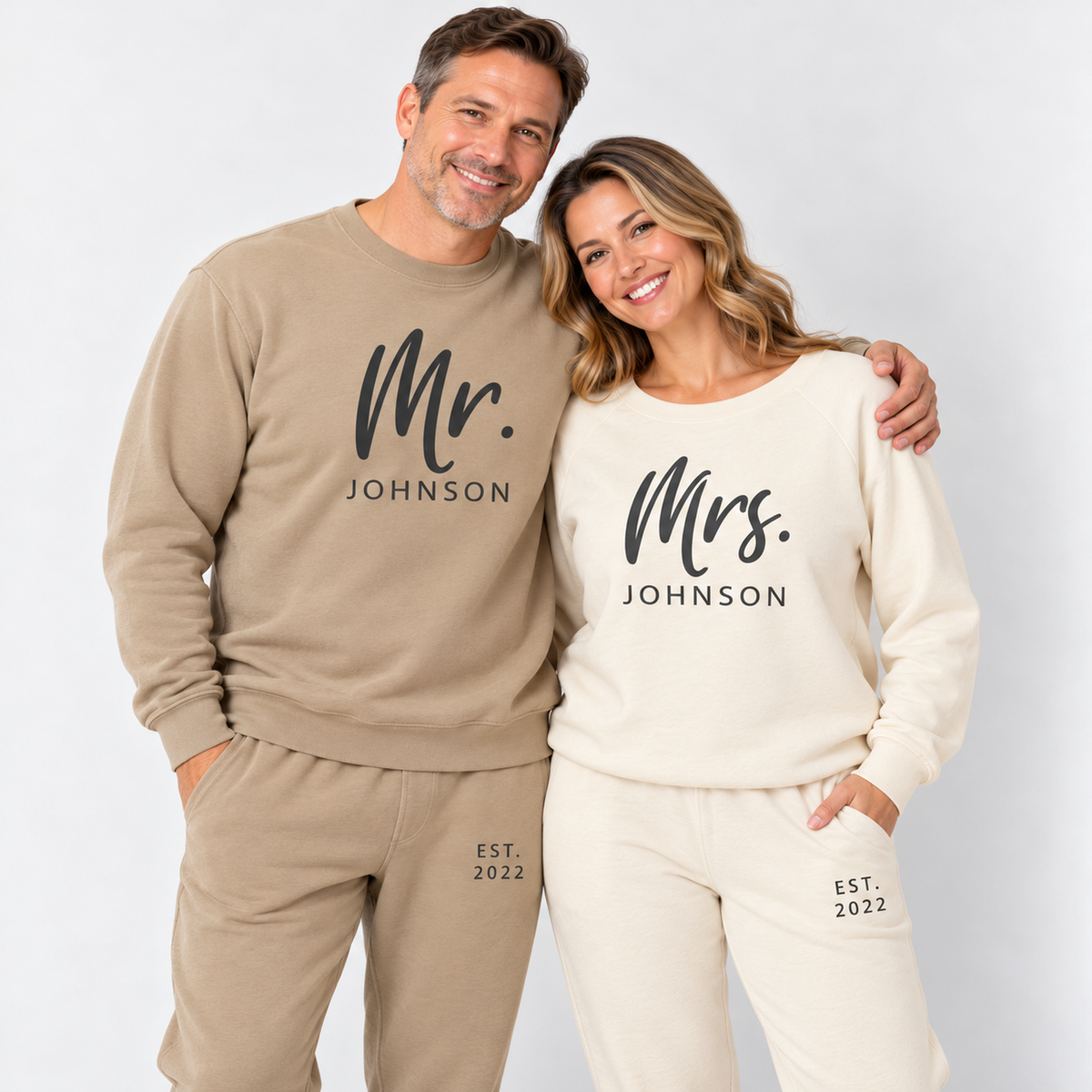 A couple wearing matching Pigment Sandstone and Bone sweatshirts and joggers personalized with Mr. Johnson and Mrs. Johnson lettering and EST. 2022 details, photographed in a clean studio.