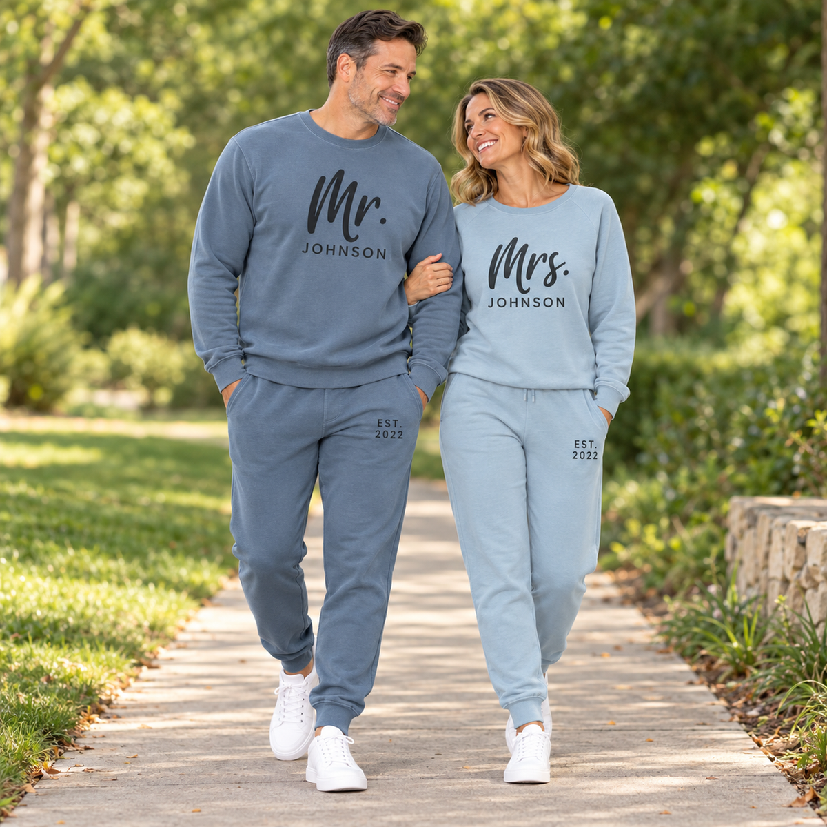 A couple walking together in a park wearing matching Pigment Slate Blue and Misty Blue sweatshirts and joggers personalized with Mr. Johnson and Mrs. Johnson lettering and EST. 2022 details.