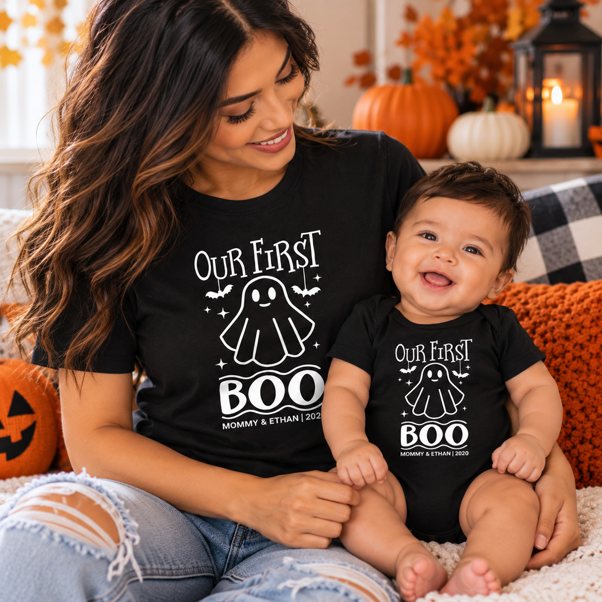 Mother and baby boy wearing matching black Our First Boo Halloween shirts with ghost and bat designs in a cozy autumn setting