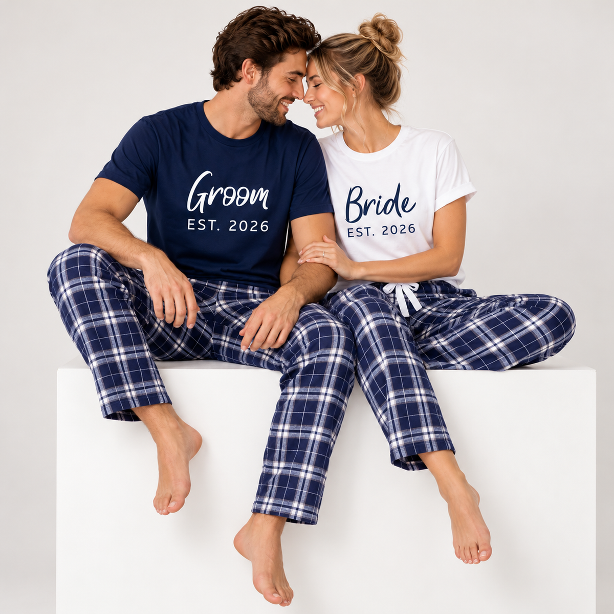 Couple wearing a navy blue Groom T-shirt and white Bride T-shirt, both with EST. 2026 prints and Navy-Silver plaid pajama pants.