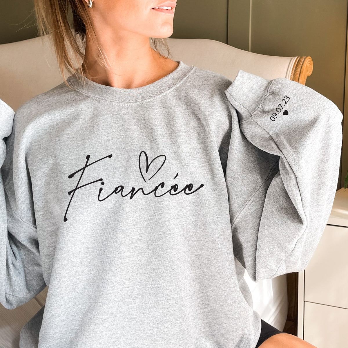 Woman wearing a Fiancée sweatshirt with elegant script lettering on the front and a personalized engagement date with a heart on the sleeve.