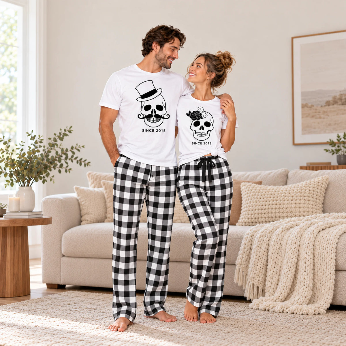 Personalized Halloween matching couple pajamas with skull T-shirts and black and white buffalo plaid flannel pants, worn by a couple in a cozy home