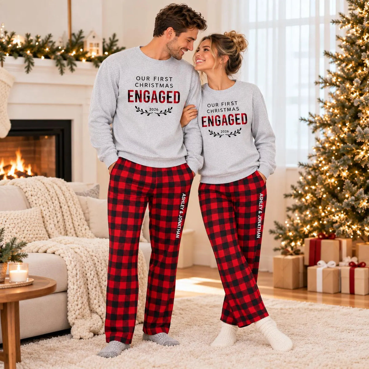 A couple wearing matching Our First Christmas Engaged 2026 sweatshirts and personalized plaid flannel pajama pants with custom names printed on the legs, posing together in a Christmas-decorated living room.