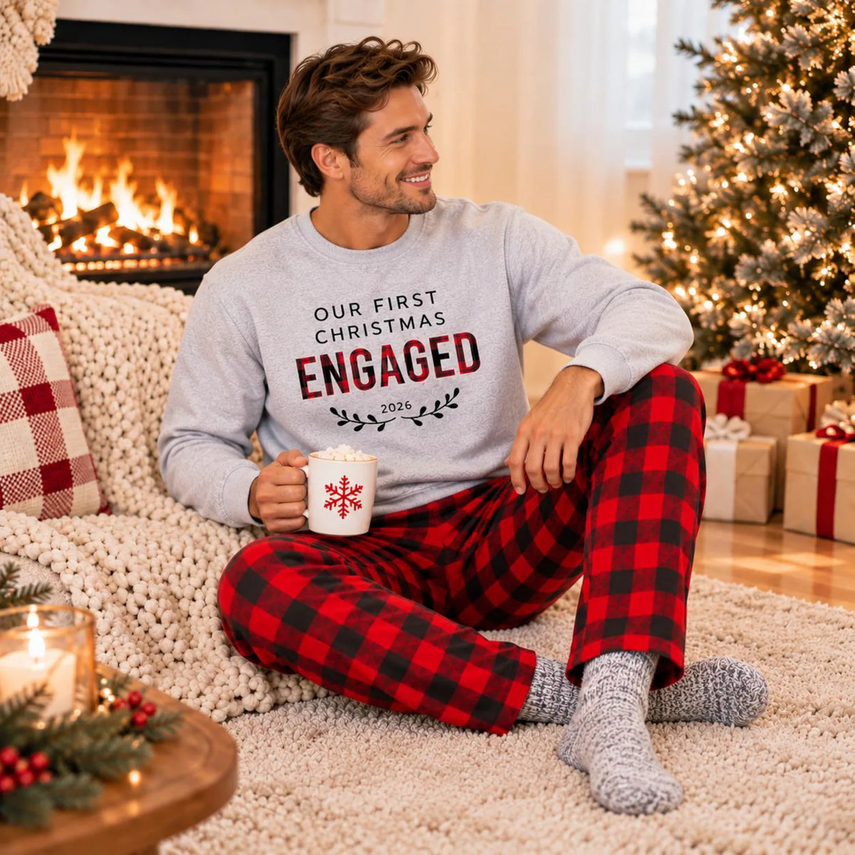 A man wearing an Our First Christmas Engaged 2026 sweatshirt and plaid flannel pajama pants, relaxing on a cozy knitted blanket beside a fireplace and decorated Christmas tree.