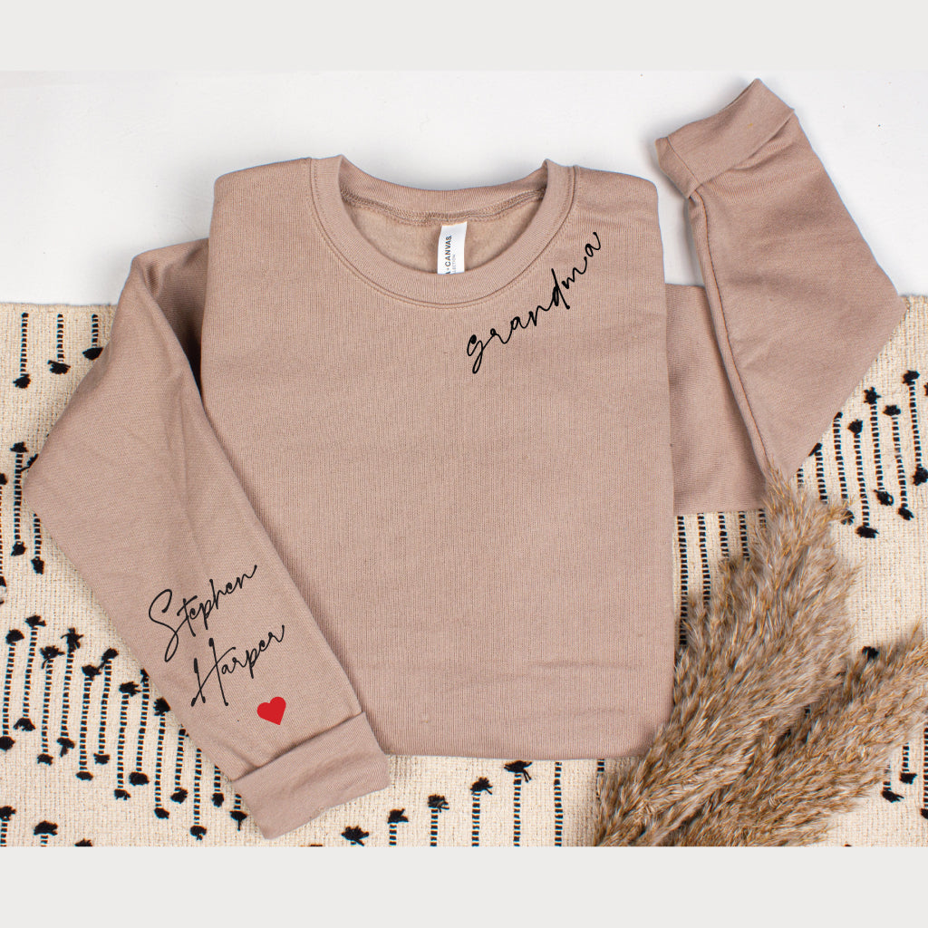Tan personalized Grandma sweatshirt with custom grandkids' names and a red heart printed on the sleeve, styled in a flat lay with a patterned blanket and dried grasses.