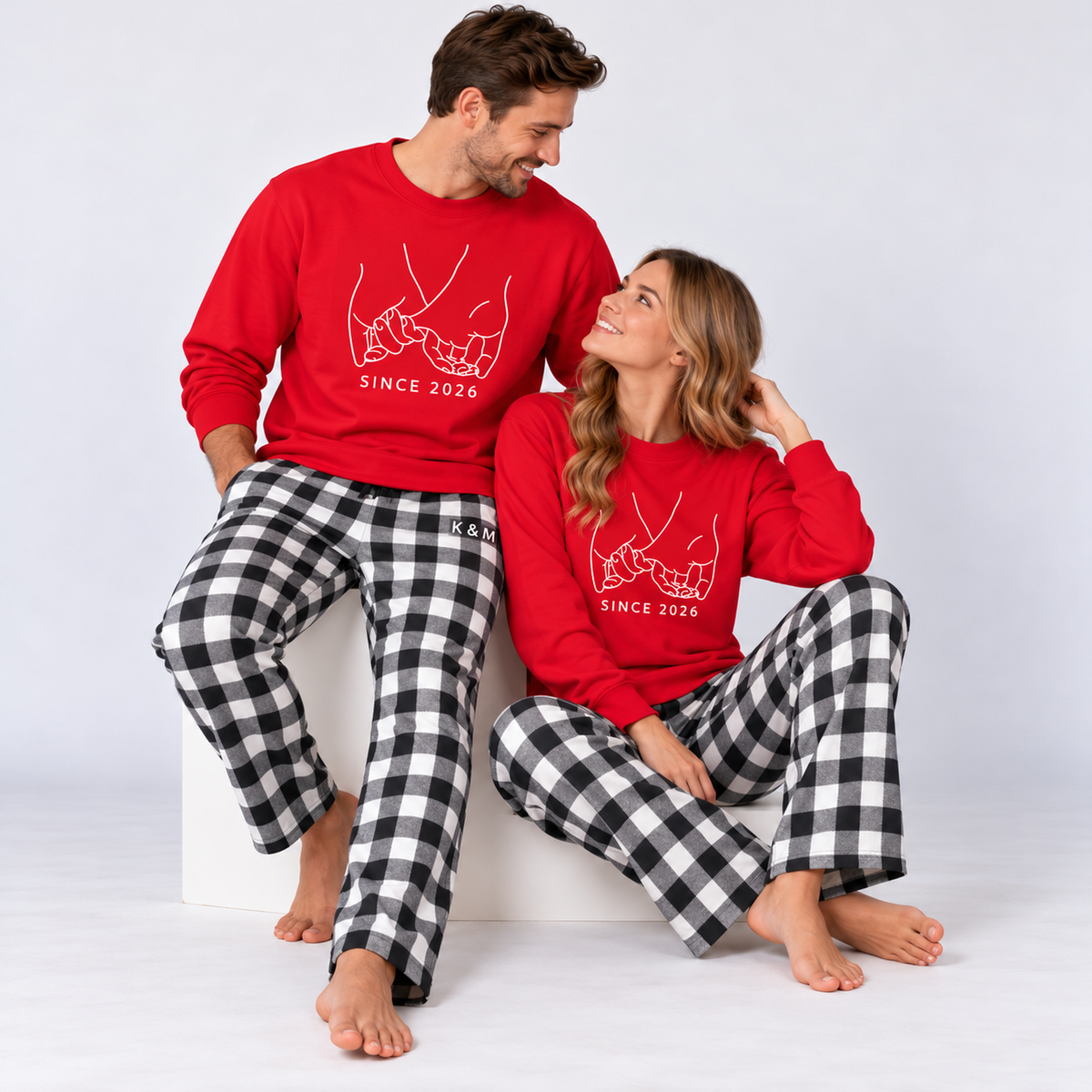 A couple wearing matching red Pinky Promise sweatshirts with the original white “SINCE 2026” hand illustration and black-and-white plaid pajama pants, featuring K & M lettering on the man’s pants.