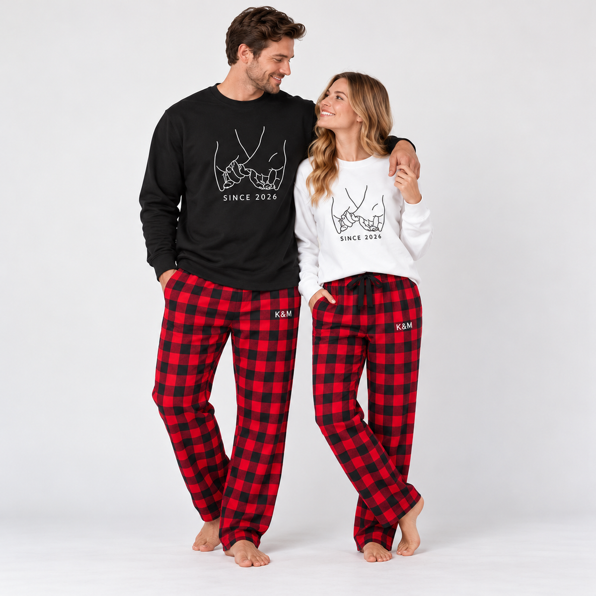 A couple wearing matching Pinky Promise sweatshirts, a black sweatshirt for him and a white sweatshirt for her, with red and black plaid pajama pants personalized with K & M lettering.