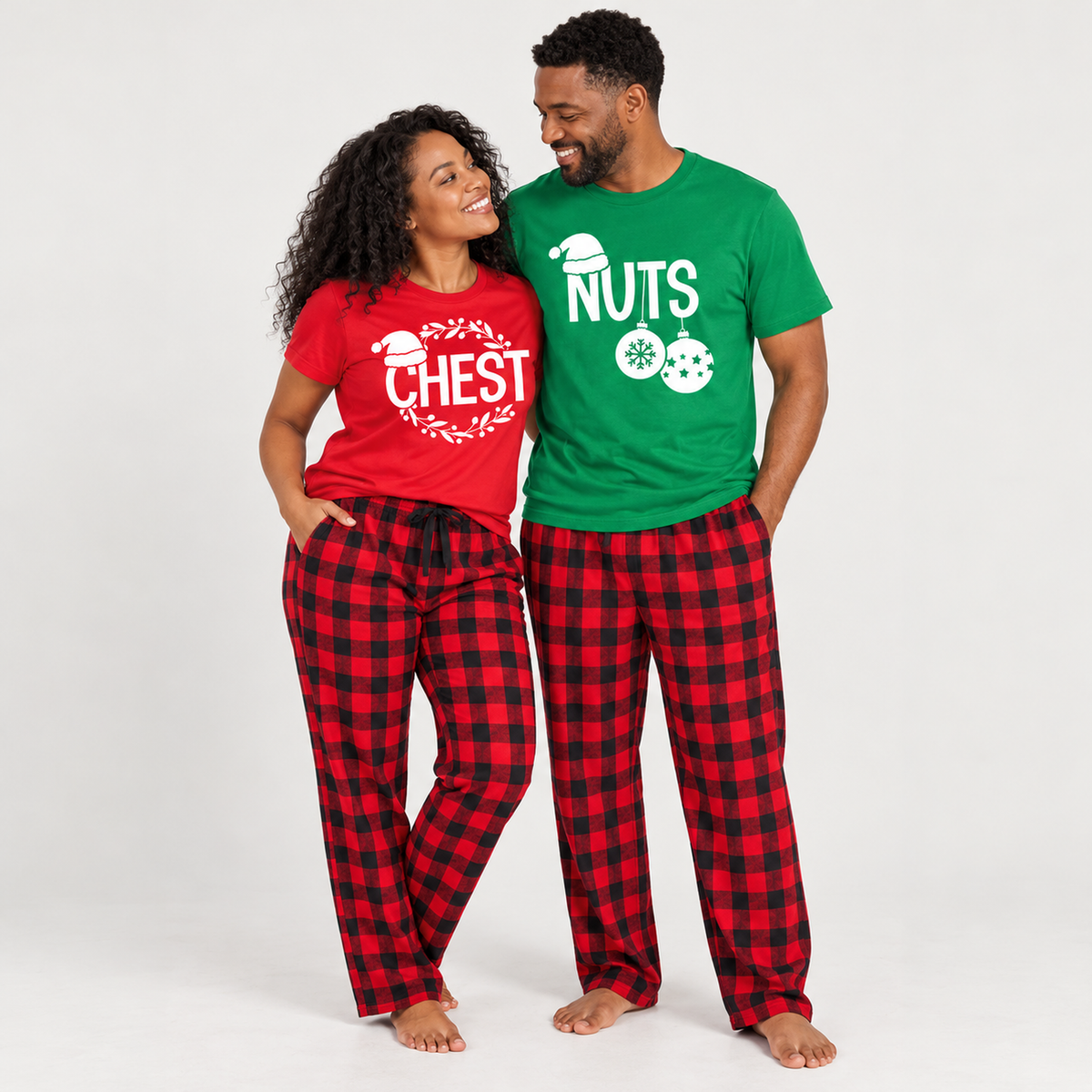 A couple wearing matching Chest and Nuts Christmas pajamas in a studio, featuring a red women's T-shirt with a white holiday wreath design, a Kelly green men's T-shirt with white Christmas ornament graphics, and matching red-and-black buffalo plaid flannel pajama pants.
