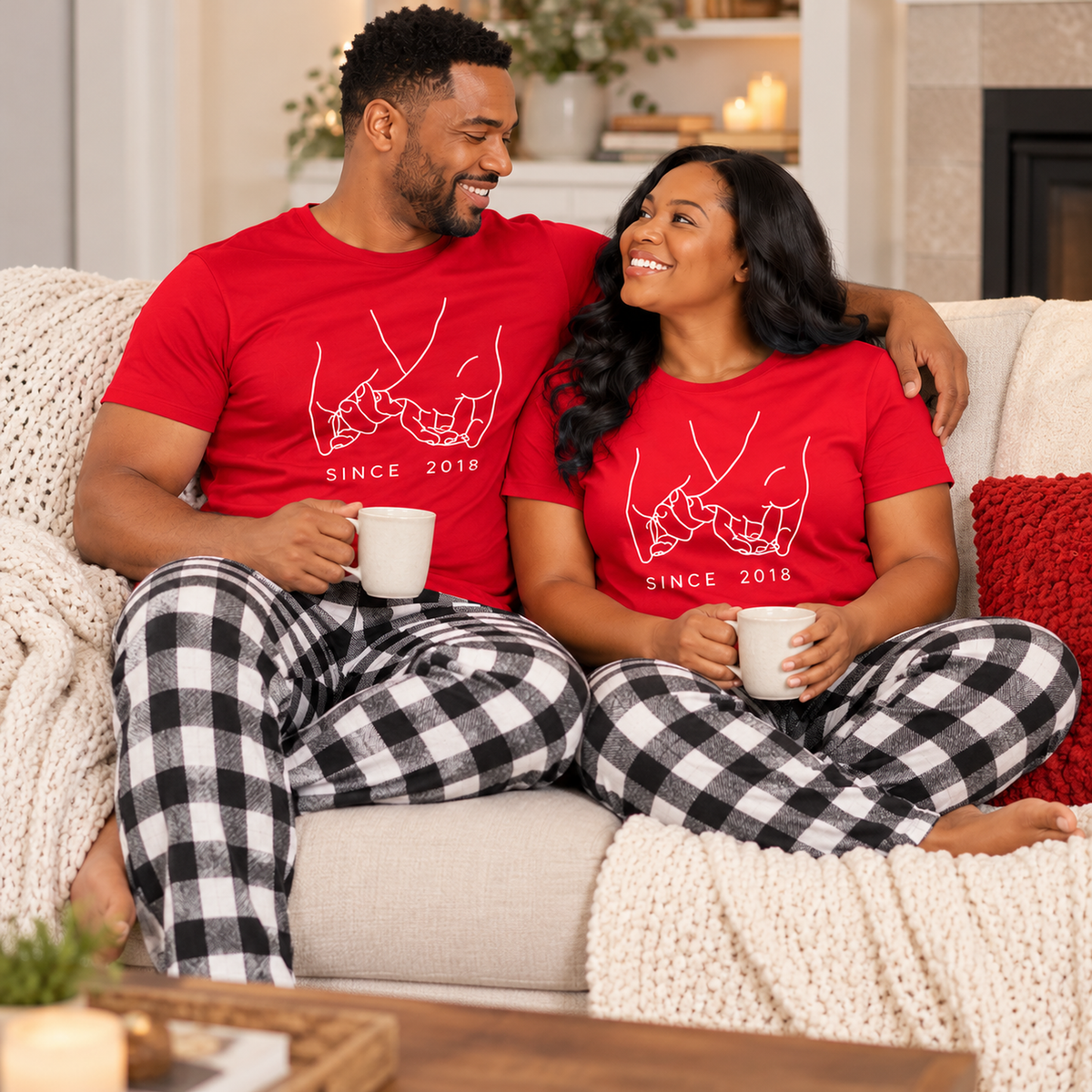 Couple relaxing together on a cozy sofa, wearing matching red T-shirts with a white holding pinkies design and custom established year, paired with black and white buffalo plaid flannel pajama pants. Warm home interior with soft blankets, pillows, and gentle ambient lighting.