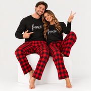 An engaged couple sitting playfully on a white studio cube, wearing matching long-sleeve jersey tees with “fiancé EST. 2027” and “fiancée EST. 2027” prints, paired with red and black buffalo plaid flannel pajama pants.