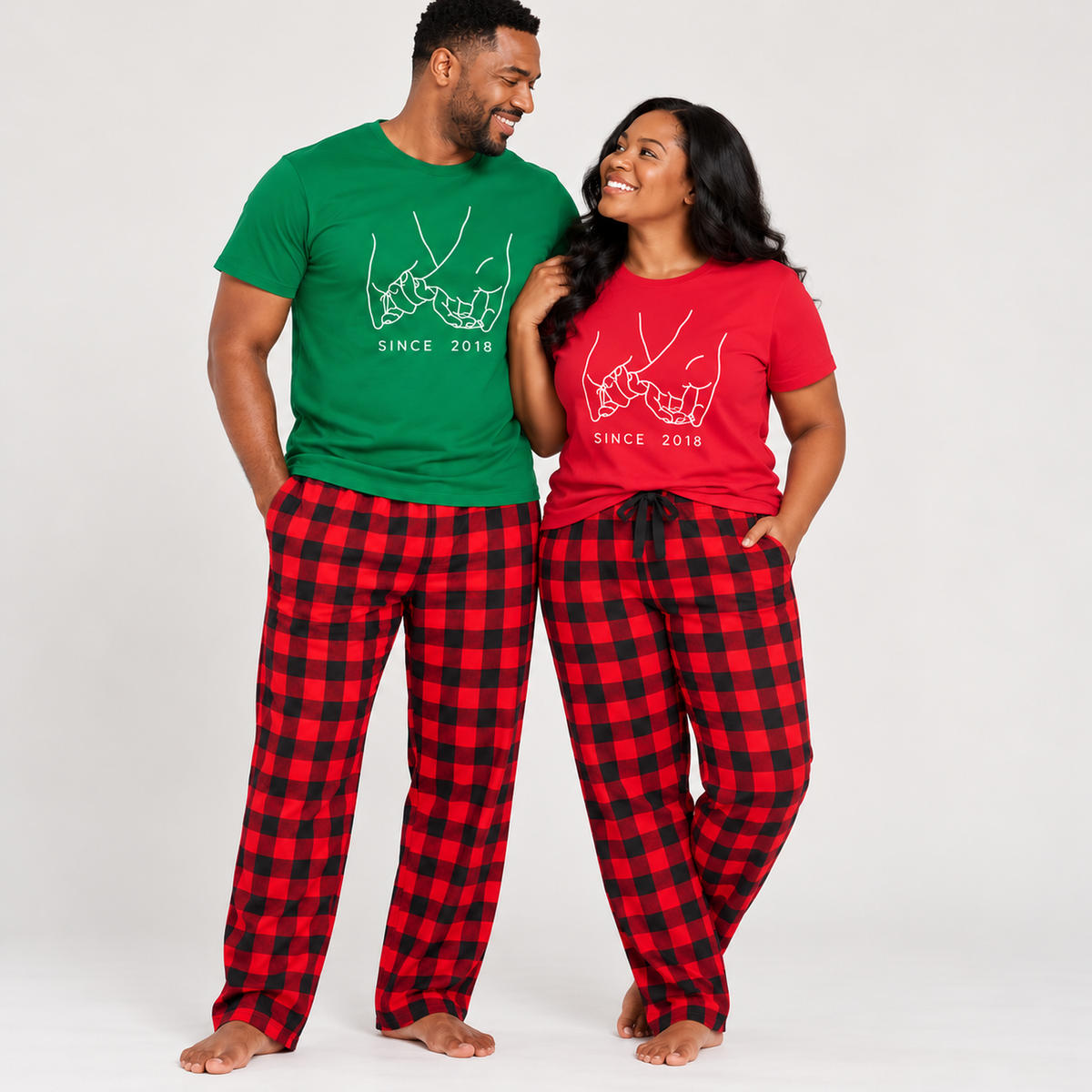 Couple wearing personalized matching pajamas featuring a Kelly green T-shirt for the man and a red T-shirt for the woman, both with a white holding pinkies illustration and custom established year, paired with red and black buffalo plaid flannel pajama pants. The woman's pajama pants feature centered black drawstring ties.