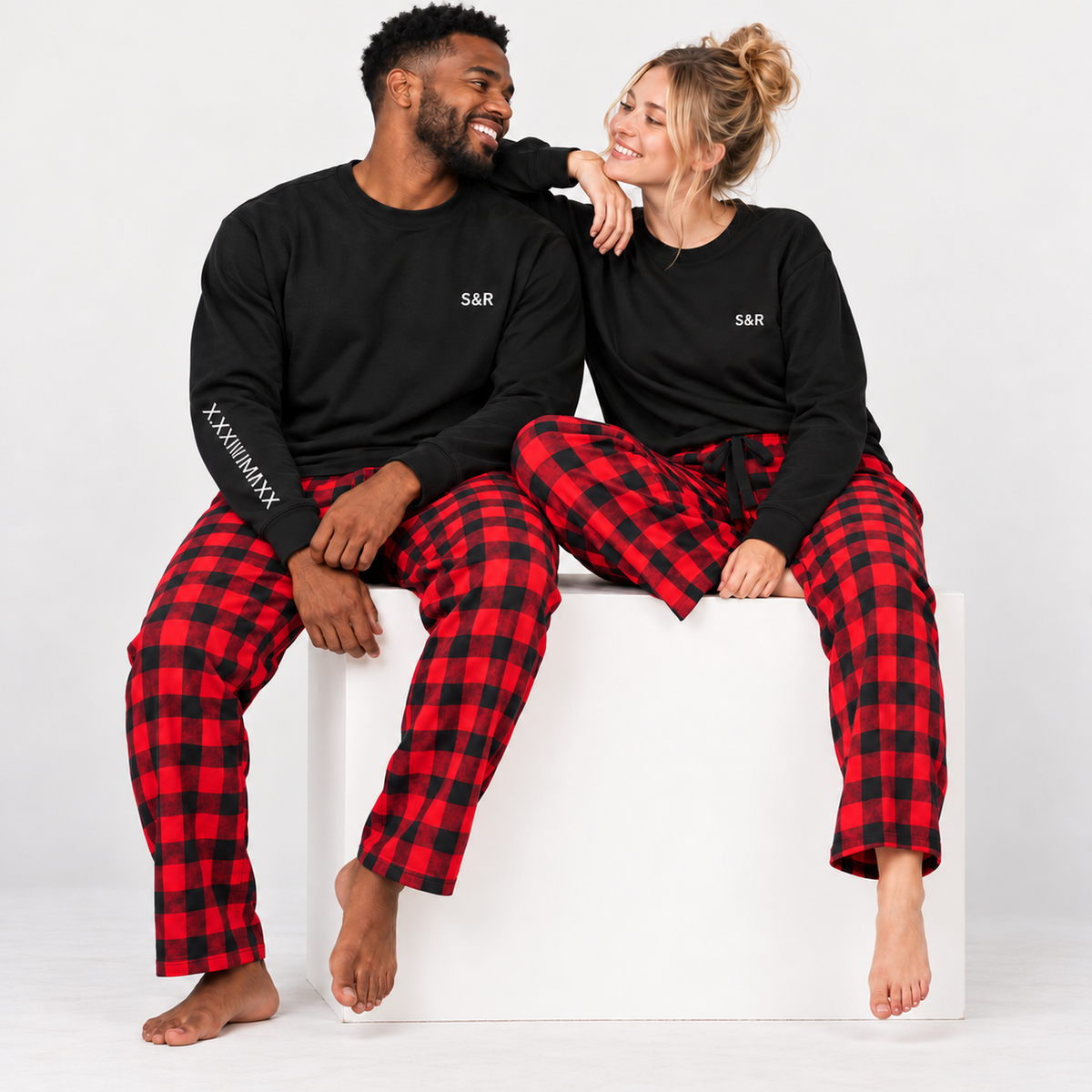 Couple wearing matching black sweatshirts with personalized S&R chest initials and Roman numeral sleeve print, paired with red and black buffalo plaid flannel pajama pants.