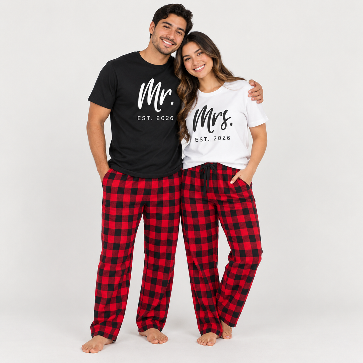 Young couple wearing matching Mr. and Mrs. T-shirts with 2026 prints and red-and-black buffalo plaid flannel pajama pants in a clean studio setting.