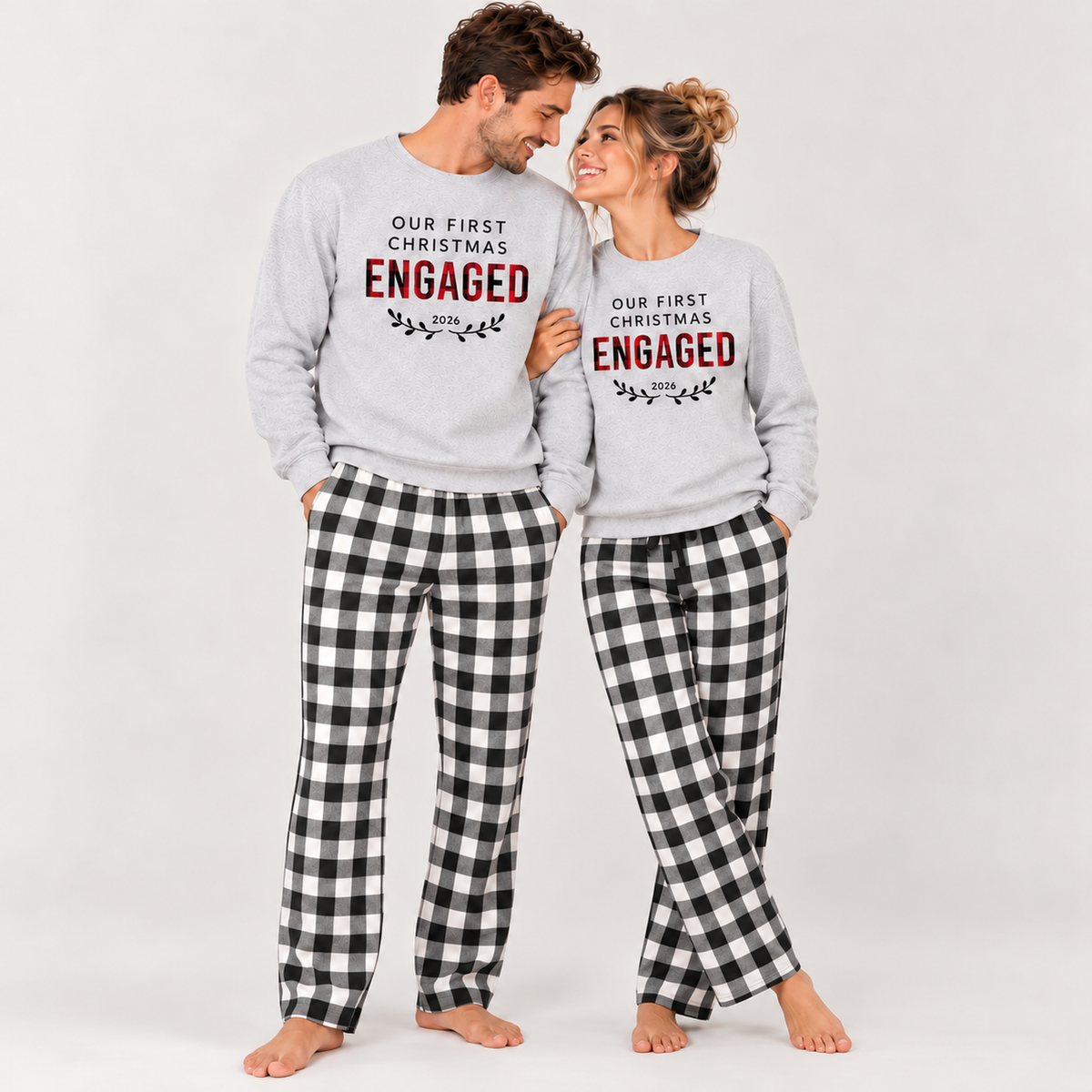 A couple wearing matching Our First Christmas Engaged 2026 sweatshirts and black-and-white plaid pajama pants, standing together in a clean studio setting.