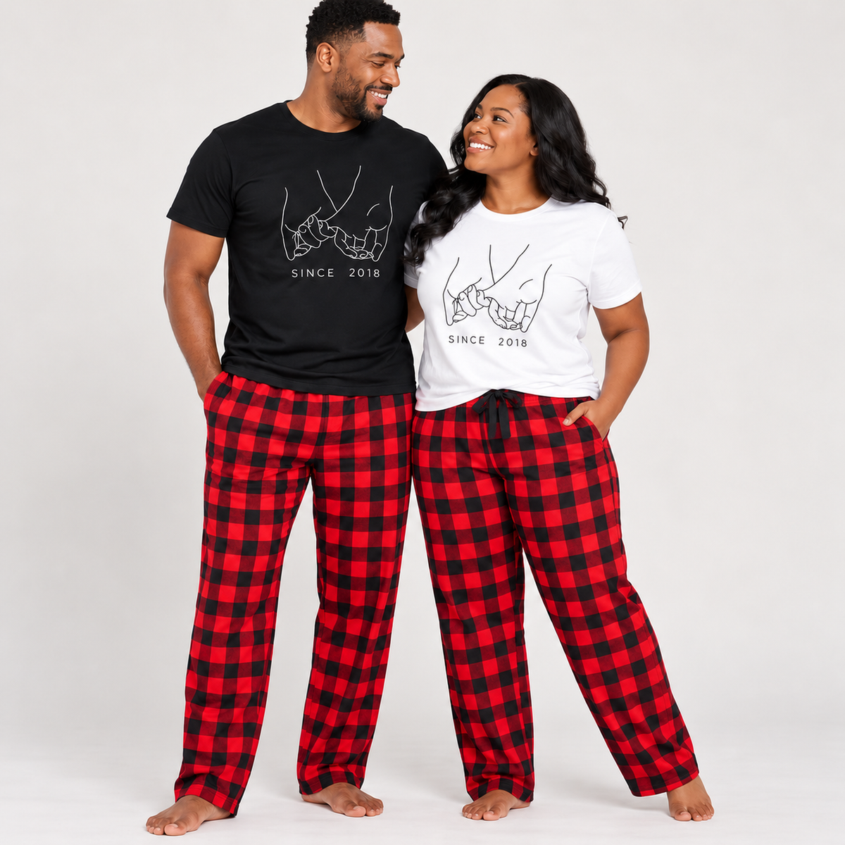 Couple wearing personalized matching pajamas featuring a black T-shirt for him and a white T-shirt for her, both with a holding pinkies design and custom established year, paired with red and black buffalo plaid flannel pajama pants. The woman’s pajama pants have centered black drawstring ties.